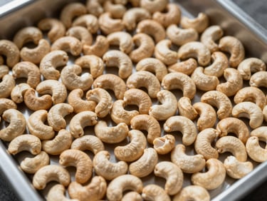 Overhead close-up of raw cashew nuts spread across a stainless sorting tray in a processing facility, natural overhead light revealing texture and color gradation