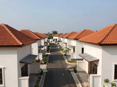 Wide daylight exterior establishing shot of a completed single-storey residential cluster in Bandung, off-white rendered walls, terracotta roof tiles, paved internal road cutting through center frame, clear midday light