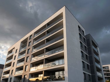 Wide-angle architectural photography of a modern apartment facade with clean anthracite concrete lines, dramatic sunset lighting reflecting off glass balconies, dark atmospheric sky.