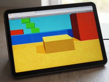 Angled screen capture of a browser game with bold colored blocks and a game-over screen, neutral desk surface visible at the bottom edge, overhead natural light
