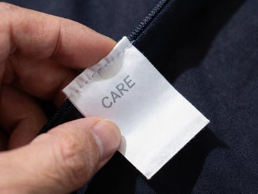 Extreme close-up of a care label being read under bright overhead daylight, fingers holding the tag flat against a dark garment, thread detail visible at the seam edge