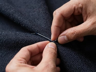 Extreme close-up of hands inspecting a seam on a dark wool garment under bright work lighting, thread and weave visible, deliberate and careful framing