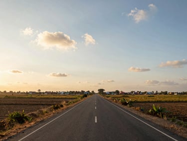 Wide-angle view of a Srikakulam district road through farmland, flat agricultural fields on both sides, a distant cluster of village buildings on the horizon, golden-hour sidelight, open sky with scattered clouds