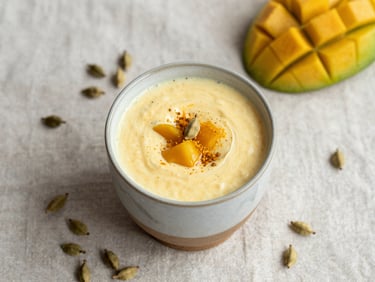 Overhead flat-lay of a mango lassi in a ceramic glass, surrounded by loose cardamom pods on a linen cloth