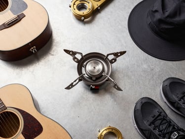 Overhead flat-lay of modern camping gear, including a sleek camp stove, brass acoustic guitar components, and technical sports gear, organized on a clean steel surface, neutral daylight, 50mm