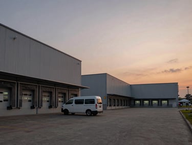 Wide shot of an Indonesian industrial estate at dusk, factory buildings with loading docks visible, a service van parked outside a facility, ambient orange-grey sky, utilitarian working environment