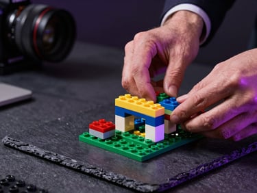 Macro shot of executive hands assembling complex structures with LEGO Serious Play bricks on a dark slate table, dramatic purple rim lighting, cinematic depth of field.