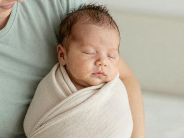 Soft editorial photography, natural morning daylight, close-up of a mother gently holding a sleeping newborn baby wrapped in a soft beige cotton blanket, shallow depth of field, 35mm