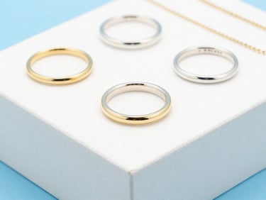 Macro shot of silver and gold rings and necklaces on a clean white display block, bright high-key studio lighting, crisp sky blue accents, 50mm lens