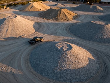 Drone overview of a gravel and sand stockpile yard in Loreto measured against a parked pickup truck for human scale, golden hour light raking across the material mounds, aggregate textures clearly visible from above