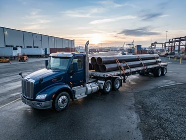 Wide-angle heroic shot of a modern MAAS TRUCKING semi-truck with a flatbed trailer loaded with industrial pipes, parked at a terminal at dawn, crisp morning daylight, high-contrast cool tones, wide-angle heroic lens, 35mm.