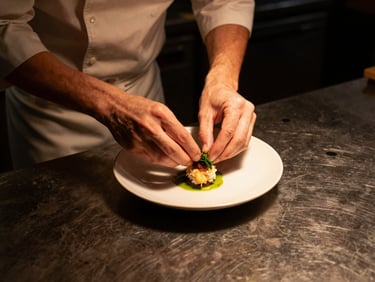 Close overhead shot of a chef's hands placing a garnish on a small plate in a dimly lit restaurant kitchen, warm amber light from above, dark worn countertop, documentary style