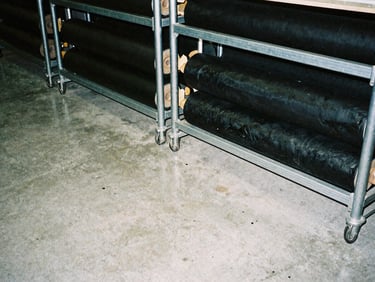 Raw concrete studio floor with industrial metal racks and heavy black garment rolls, direct harsh flash lighting, high contrast, desaturated tones, 35mm film