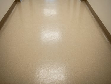 Close-up overhead study of quartz epoxy floor in a healthcare facility corridor, even warm light, clean surface texture visible with subtle aggregate pattern, no people, wall base trim visible at frame edge
