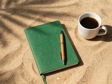A flat-lay of a green notebook, a wooden pen, and a cup of coffee on a warm sand surface with soft morning light.
