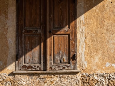 Close-up of a weathered wooden shutter half-open on a stone wall, afternoon light casting a diagonal shadow across faded ochre plaster, eye-level framing, texture of aging wood grain visible