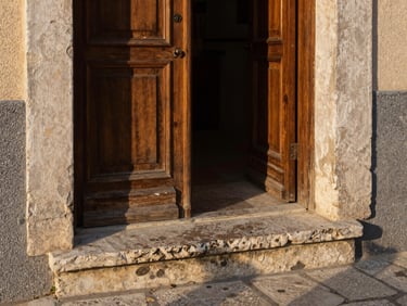 Eye-level close-up of a worn stone threshold and an open wooden door in a Mediterranean village, late afternoon light raking across the surface revealing texture and age, no people, generous shadow in the doorway interior
