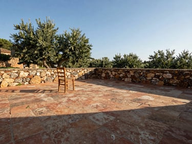 Wide environmental shot of an empty stone farmhouse terrace in the Costa Brava, morning light casting long shadows across weathered terracotta tiles, a single wooden chair facing an olive grove, no people, eye-level framing