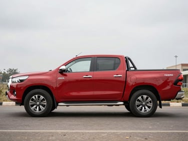 Full side view of a red Toyota Hilux pickup on a neutral street-level Accra background, flat overcast light, load bed and cabin both visible