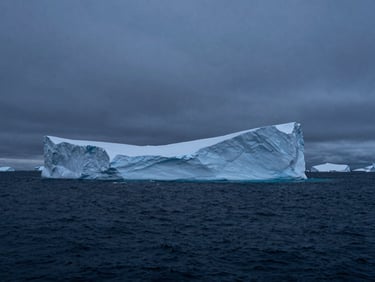 Wide cinematic shot of a massive, sharp-edged monolithic iceberg floating in a dark, quiet arctic ocean under a cold overcast sky, deep blue-hour shadows, high contrast.