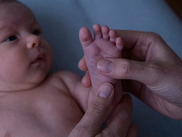 A close-up of a mother's hand gently holding a tiny newborn foot, warm dim lighting, soft twilight tones, shallow depth of field, 35mm