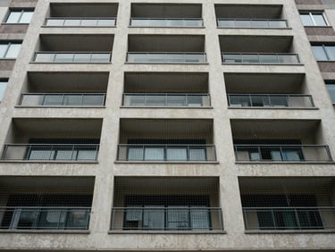 Exterior facade of a commercial building or housing society, invisible grille netting installed across multiple floors of balconies, overcast mid-day light, wide architectural shot from ground level looking up, grille barely visible against the building surface