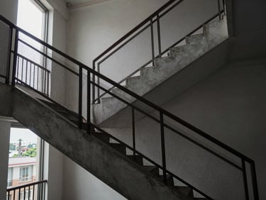 Interior staircase of a mid-rise residential building, invisible netting installed along the open railing side, overcast daylight from a nearby window, wide environmental shot showing the full vertical run of stairs, grille barely perceptible