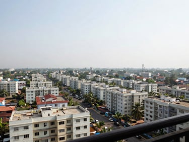 Wide panoramic view of a Vasai or Virar residential neighbourhood from a high balcony, rows of apartment buildings stretching toward the horizon under a bright overcast sky, an invisible grill netting barely visible in the foreground frame, natural daylight, environmental and unposed