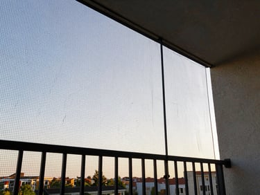 Inside view of a residential apartment balcony, near-invisible netting strung between posts, bright afternoon sky visible beyond, no obstructing bars, natural daylight, wide environmental framing from inside looking out