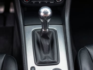 Close-up technical shot of a gated manual shifter in a classic sports car, brushed aluminum console, clean leather boot, straight-on eye-level framing.