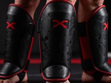 Close-up of black MMA shin guards with red accents, showing multi-layered padding, dramatic lighting.