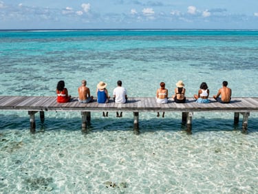 A wide environmental shot of a group of relaxed travelers on a wooden dock over crystal-clear water