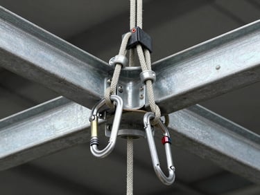 Close-up overhead framing of a rope access anchor point on a steel beam, double carabiners locked and loaded, kermantle rope running taut across the frame, studio-bright industrial lighting revealing texture of every component, no human visible — only the rigging hardware in precise detail