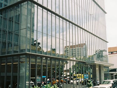 Sleek modern office building facade with reflective glass, showing a glimpse of a bustling Indonesian street, bright natural daylight, soft shadows, 35mm.