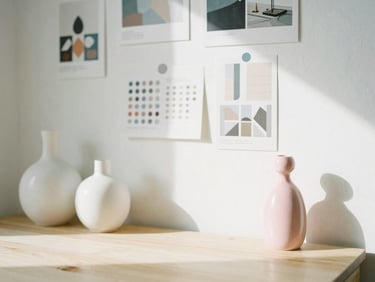 Bright sun-drenched workspace with ceramic vases, design mood boards on a white wall, and a pink ceramic figurine on a wooden desk, soft shadows, 35mm