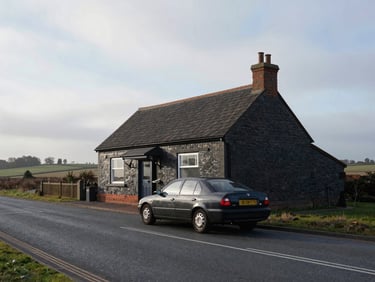 Wide documentary shot of a dark saloon car on a quiet North Lincolnshire town road, overcast morning light, no pedestrians, flat landscape visible in background