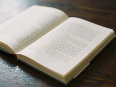 An open hardcover book on a dark wooden table, showing elegant serif typography on textured cream paper, soft side lighting, shallow depth of field.