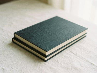 A stack of two minimalist books with textured charcoal covers, lying on a raw linen surface under soft natural light.