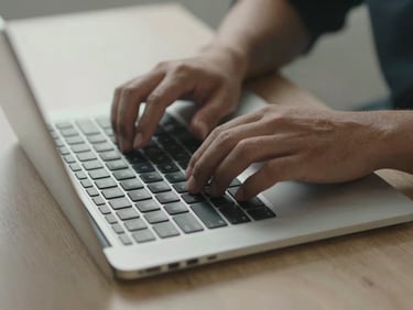 Close-up overhead of hands typing on a laptop keyboard on a plain office desk, soft daylight from the left, no screen content visible, shallow depth, neutral background