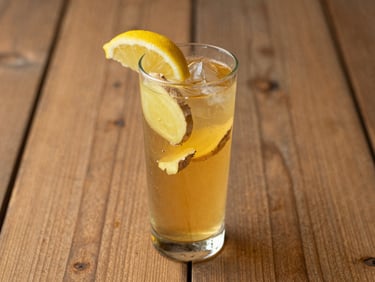 Tall glass of golden ginger brew with fresh ginger slices and a lemon wedge, rustic wooden surface, soft lighting.