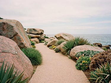 Private garden path with coastal plants and pink granite boulders, looking towards the sea under soft overcast Breton sky, warm editorial photography