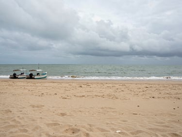 Panoramic overcast-light shot of Nagaon beach, wide flat sand in the foreground, grey-green sea meeting a low cloudy horizon, a pair of fishing catamarans pulled ashore on the left edge, no tourists visible