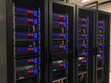 Wide shot of an AV equipment server rack in a dim technical room, indicator lights glowing blue and red, cables bundled and labelled, task lighting from below illuminating the rack face