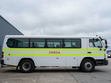 Documentary photography of a marked OMEGA QSE fleet vehicle parked outside the Watertown headquarters under overcast sky, sharp focus, industrial setting.