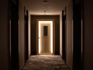 Wide interior corridor of a five-star hotel at night, a single illuminated modular pod positioned at the corridor's end as a destination focal point, brushed gold hardware catching dramatic side-lighting, minimal guests, deep shadow framing the composition