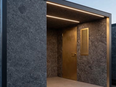 Architectural close-up of a modular wellness pod interior at a wellness center, textured obsidian wall panels and brushed gold ventilation details under cool-to-warm gradient lighting, empty and serene, spatial proportion emphasized