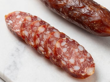 Close-up top-down view of sliced cured sausage and lomo on white marble, visible fat and seasoning detail, soft diffused daylight, no hands
