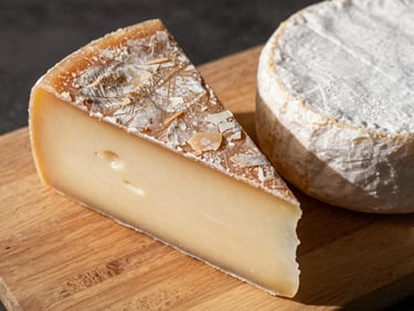 Overhead close-up of a wedge of aged manchego and a round of soft cheese resting on a bare wood board, morning side light, crumbled rind visible, no people