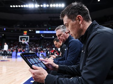 A scout in a dark jacket reviewing player film on a tablet at courtside, broadcast arena lights above, court surface visible below, other coaches in soft focus behind, intense concentration on the screen