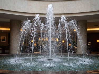 Environmental wide shot of an architectural water fountain installed in a hotel lobby interior, water jets in motion, natural and ambient light, surrounding architecture visible, no people, documentary framing
